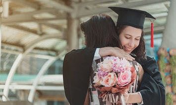 A graduate in a cap and gown hugs another person while holding a bouquet of flowers. They are outdoors, possibly at a university or event venue with a glass and metal structure.