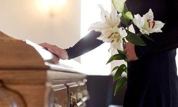 A person in black holds white lilies, resting a hand on a closed wooden casket; soft lighting and a blurred background set a solemn, respectful atmosphere.